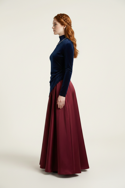 A woman in side profile wearing a dark navy velvet mock-neck top and a long, flowing burgundy satin skirt, standing against a plain light beige studio background.