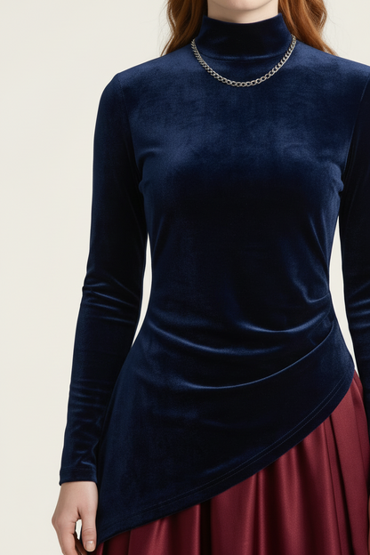 A close-up of a model wearing a navy blue velvet mock neck long-sleeve top with an asymmetrical draped hem, paired with a burgundy skirt and a silver chain necklace.
