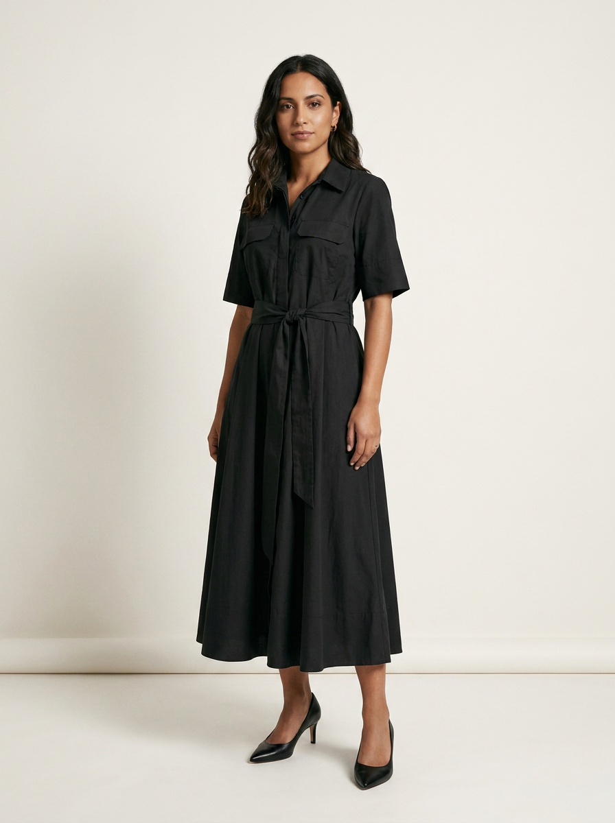 A woman modeling a black short-sleeve midi shirt dress with a tie waist belt, standing against a plain off-white studio background.