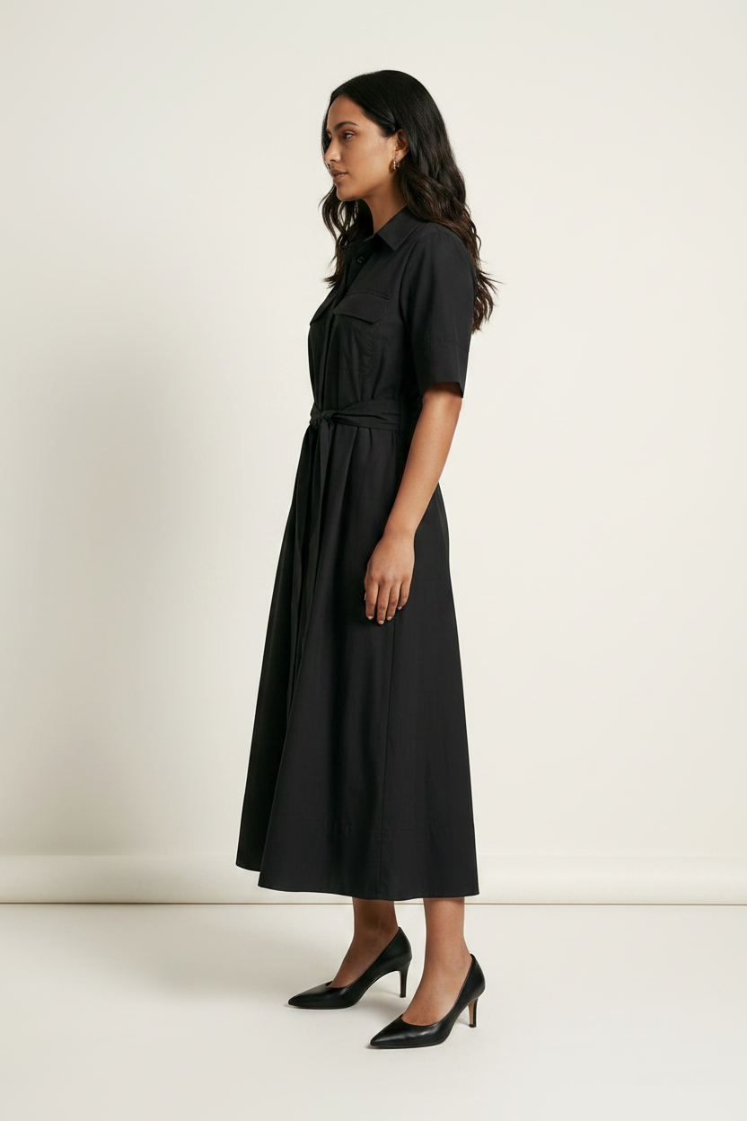 A woman modeling a black midi shirt dress with short sleeves, a collar, and a matching tie belt, paired with black pointed-toe heels, against a plain off-white studio background.