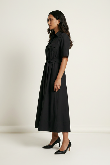 A woman modeling a black midi shirt dress with short sleeves, a collar, and a matching tie belt, paired with black pointed-toe heels, against a plain off-white studio background.