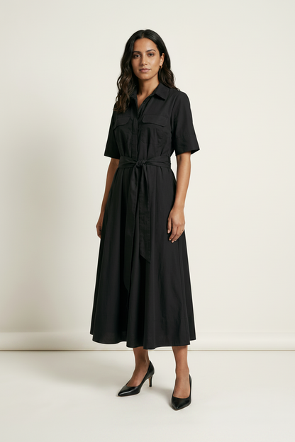 A woman modeling a black short-sleeve midi shirt dress with a tie waist belt, standing against a plain off-white studio background.