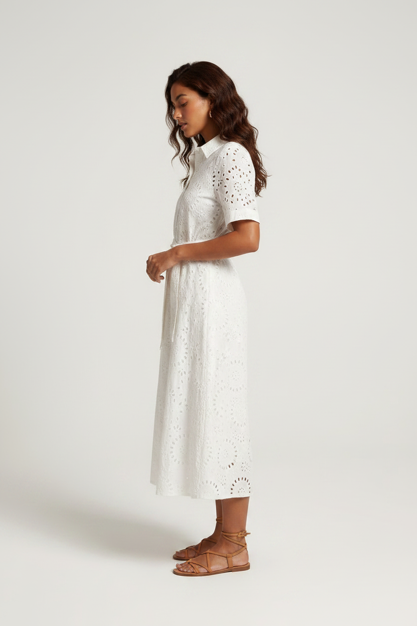 A woman modeling a white short-sleeve eyelet midi dress with a tie waist, paired with brown strappy sandals, against a light gray studio background.