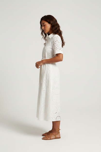 A woman modeling a white short-sleeve eyelet midi dress with a tie waist, paired with brown strappy sandals, against a light gray studio background.
