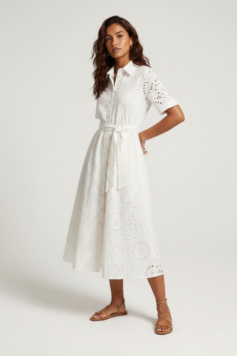 A woman modeling a white short-sleeve eyelet midi dress with a tie waist, paired with brown strappy sandals, against a light gray studio background.