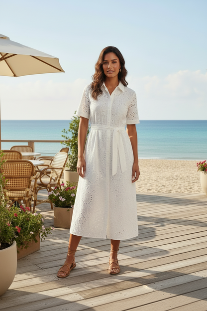 A woman stands on a wooden beachside deck wearing a white eyelet midi dress with short sleeves and tan flat sandals, with the ocean, sand, patio furniture, and umbrellas in the background.