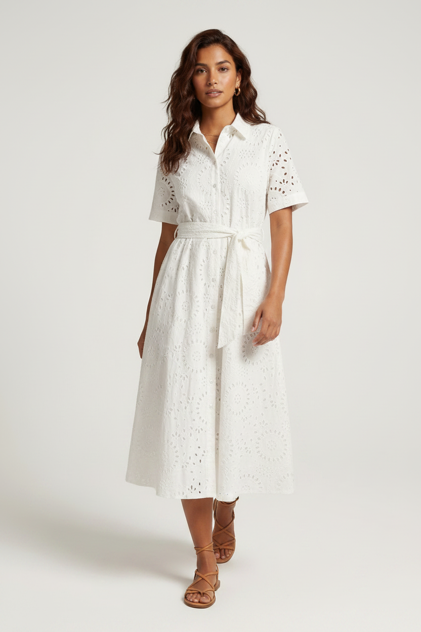 A woman modeling a white short-sleeve eyelet midi dress with a tie waist, paired with brown strappy sandals, against a light gray studio background.