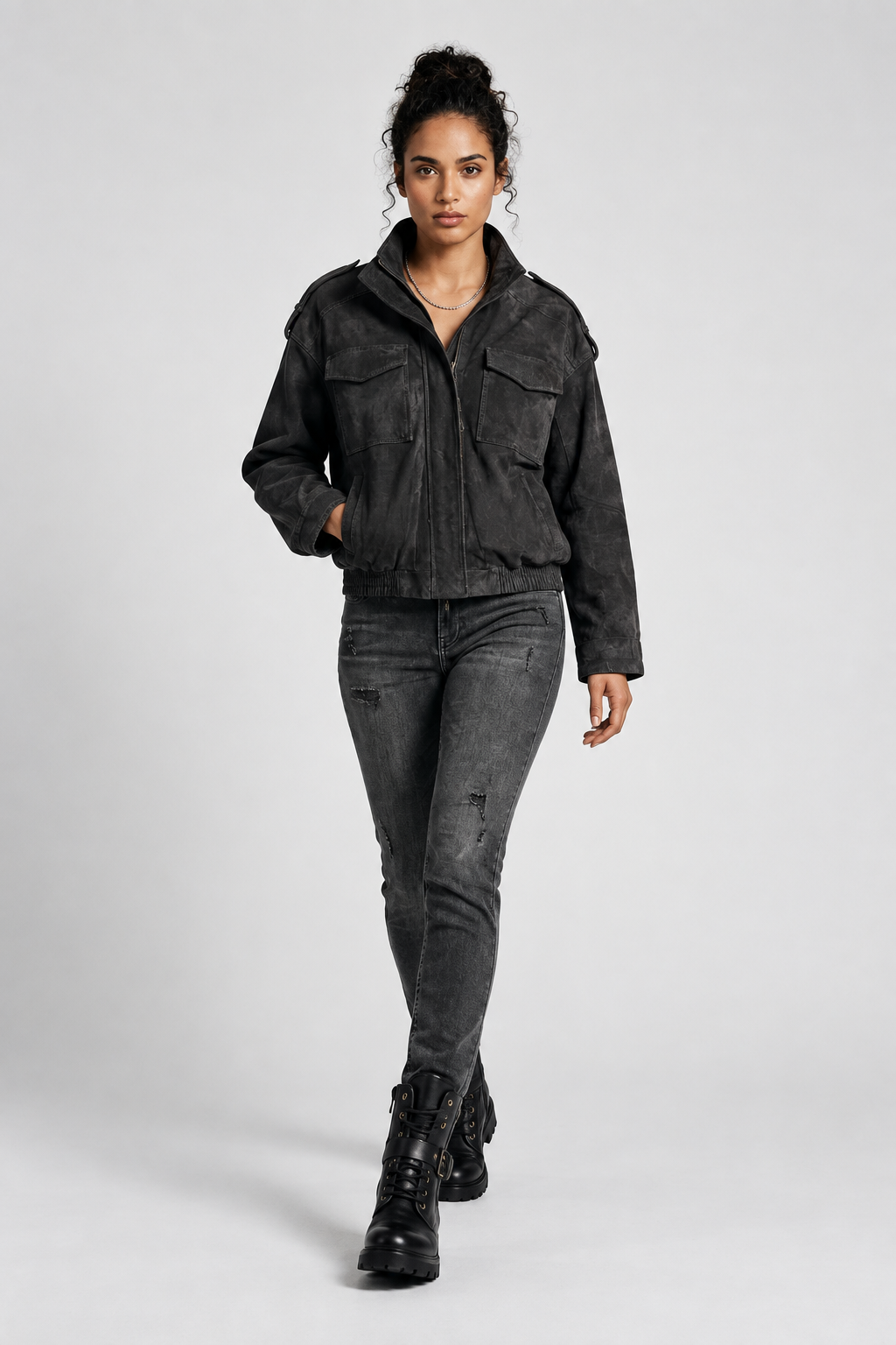 A model wearing a washed black cropped utility jacket, dark gray skinny jeans, and black combat boots, standing against a light gray studio background.