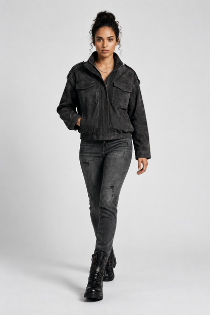 A model wearing a washed black cropped utility jacket, dark gray skinny jeans, and black combat boots, standing against a light gray studio background.