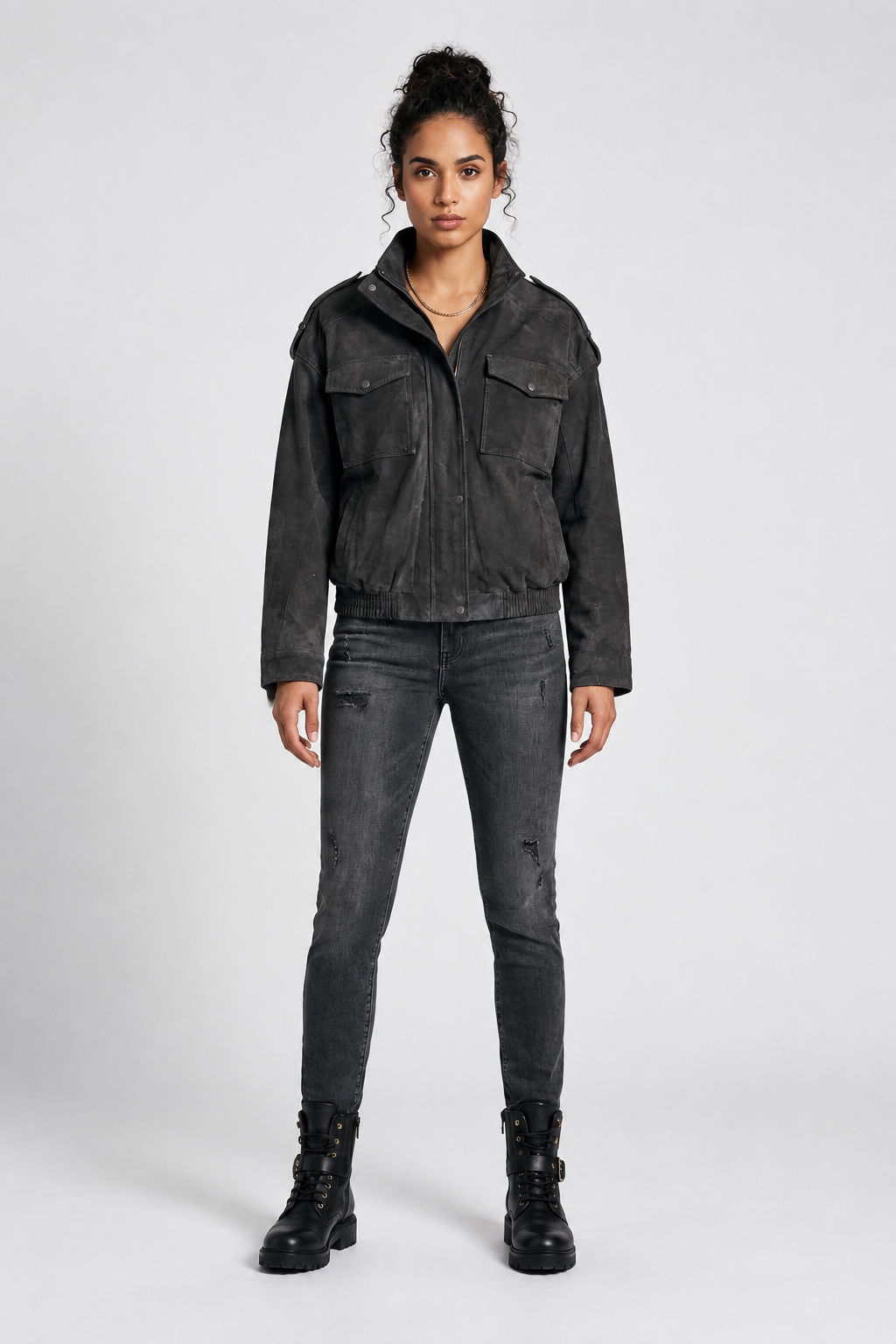 A model stands against a light gray studio background wearing a washed black cropped utility jacket, dark gray distressed skinny jeans, and black combat boots.