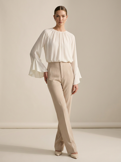 A woman modeling a white long-sleeve blouse with dramatic flared bell sleeves and beige high-waisted trousers, standing against a warm beige studio backdrop.