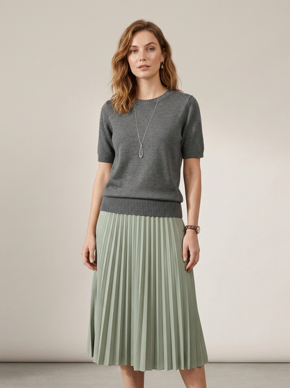 A woman stands against a plain beige studio background wearing a short-sleeve gray knit sweater and a pleated sage green midi skirt. She is also wearing a long silver pendant necklace and a watch.