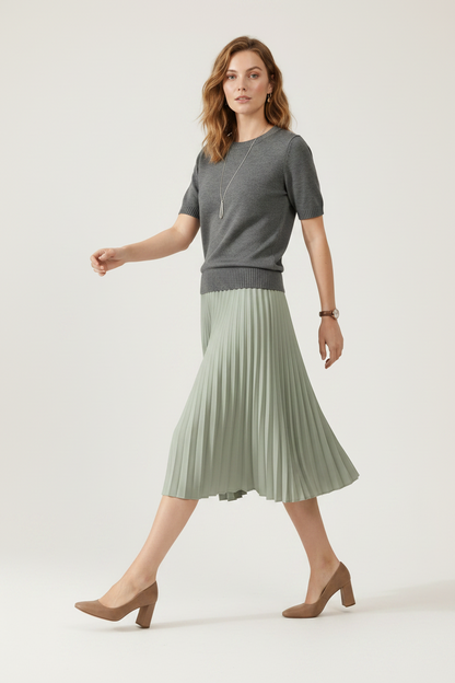 A woman stands against a plain beige studio background wearing a short-sleeve gray knit sweater and a pleated sage green midi skirt. She is also wearing a long silver pendant necklace and a watch.