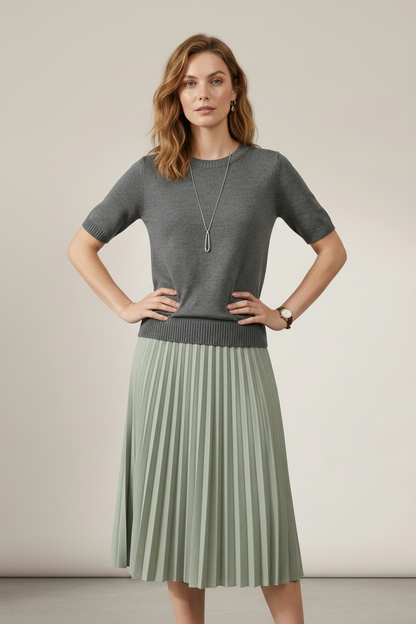 A woman stands against a plain beige studio background wearing a short-sleeve gray knit sweater and a pleated sage green midi skirt. She is also wearing a long silver pendant necklace and a watch.