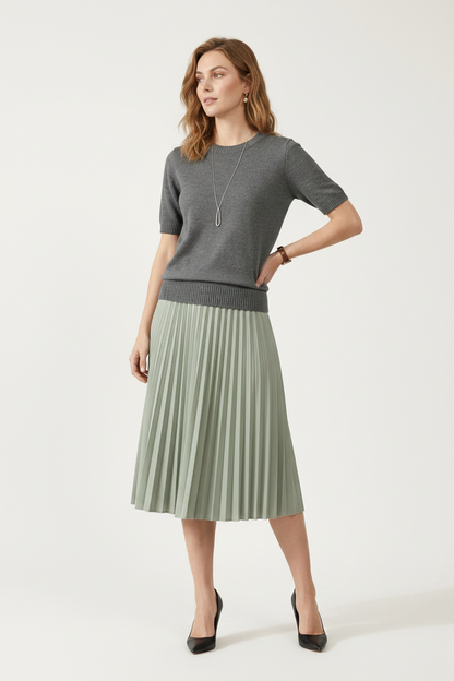 A woman stands against a plain beige studio background wearing a short-sleeve gray knit sweater and a pleated sage green midi skirt. She is also wearing a long silver pendant necklace and a watch.