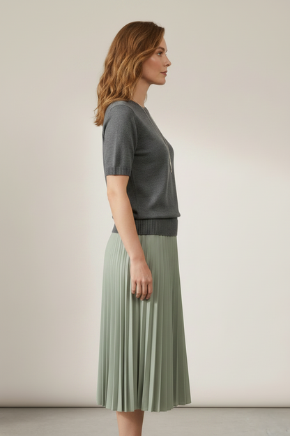 A woman stands against a plain beige studio background wearing a short-sleeve gray knit sweater and a pleated sage green midi skirt. She is also wearing a long silver pendant necklace and a watch.