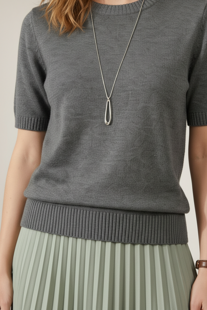 A woman stands against a plain beige studio background wearing a short-sleeve gray knit sweater and a pleated sage green midi skirt. She is also wearing a long silver pendant necklace and a watch.