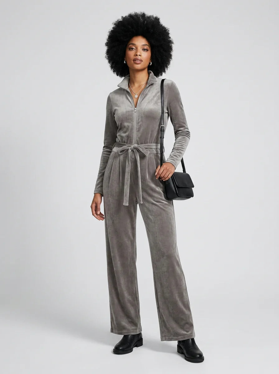 A model with a large black afro wears a gray velvet long-sleeve jumpsuit with a tie waist, paired with black boots and carrying a small black shoulder bag, against a light gray studio background.