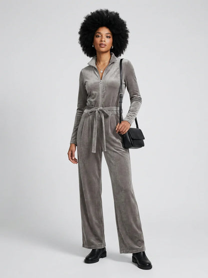 A model with a large black afro wears a gray velvet long-sleeve jumpsuit with a tie waist, paired with black boots and carrying a small black shoulder bag, against a light gray studio background.