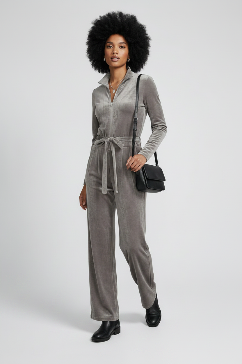 A model with an afro hairstyle wearing a gray velvet long-sleeve jumpsuit with a tie waist, black boots, and carrying a black crossbody bag against a light gray studio background.