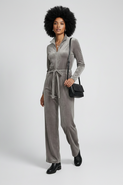 A model with an afro hairstyle wearing a gray velvet long-sleeve jumpsuit with a tie waist, black boots, and carrying a black crossbody bag against a light gray studio background.