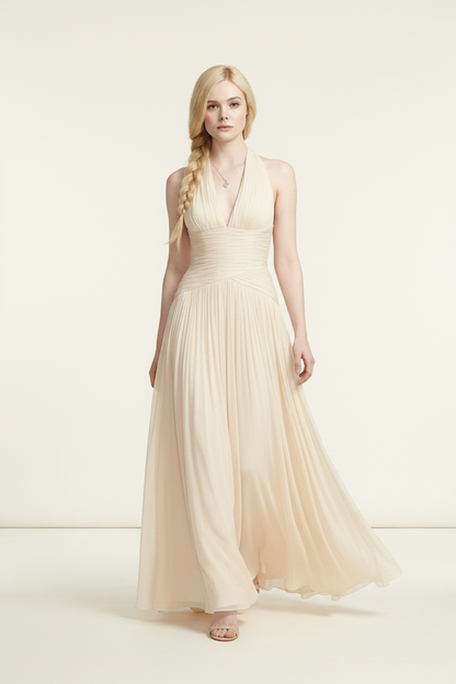 A blonde model wearing a floor-length cream halter gown with a ruched bodice and flowing tulle skirt, standing against a plain cream studio background.