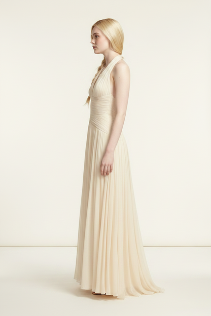 A blonde model wearing a floor-length cream halter gown with a ruched bodice and flowing tulle skirt, standing against a plain cream studio background.