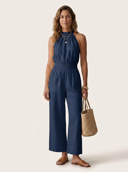 A woman modeling a navy blue halter-neck wide-leg jumpsuit, styled with layered gold necklaces, bracelets, and brown flat sandals, while holding a woven straw tote bag against a solid light beige background.