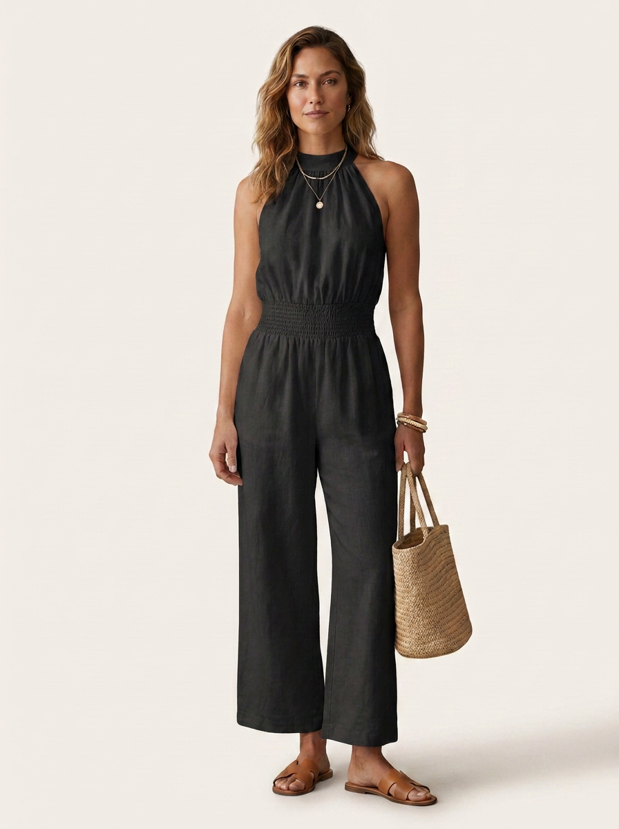 A woman modeling a black sleeveless wide-leg jumpsuit with a smocked waist, paired with a woven straw tote bag and gold jewelry, set against a plain light beige background.