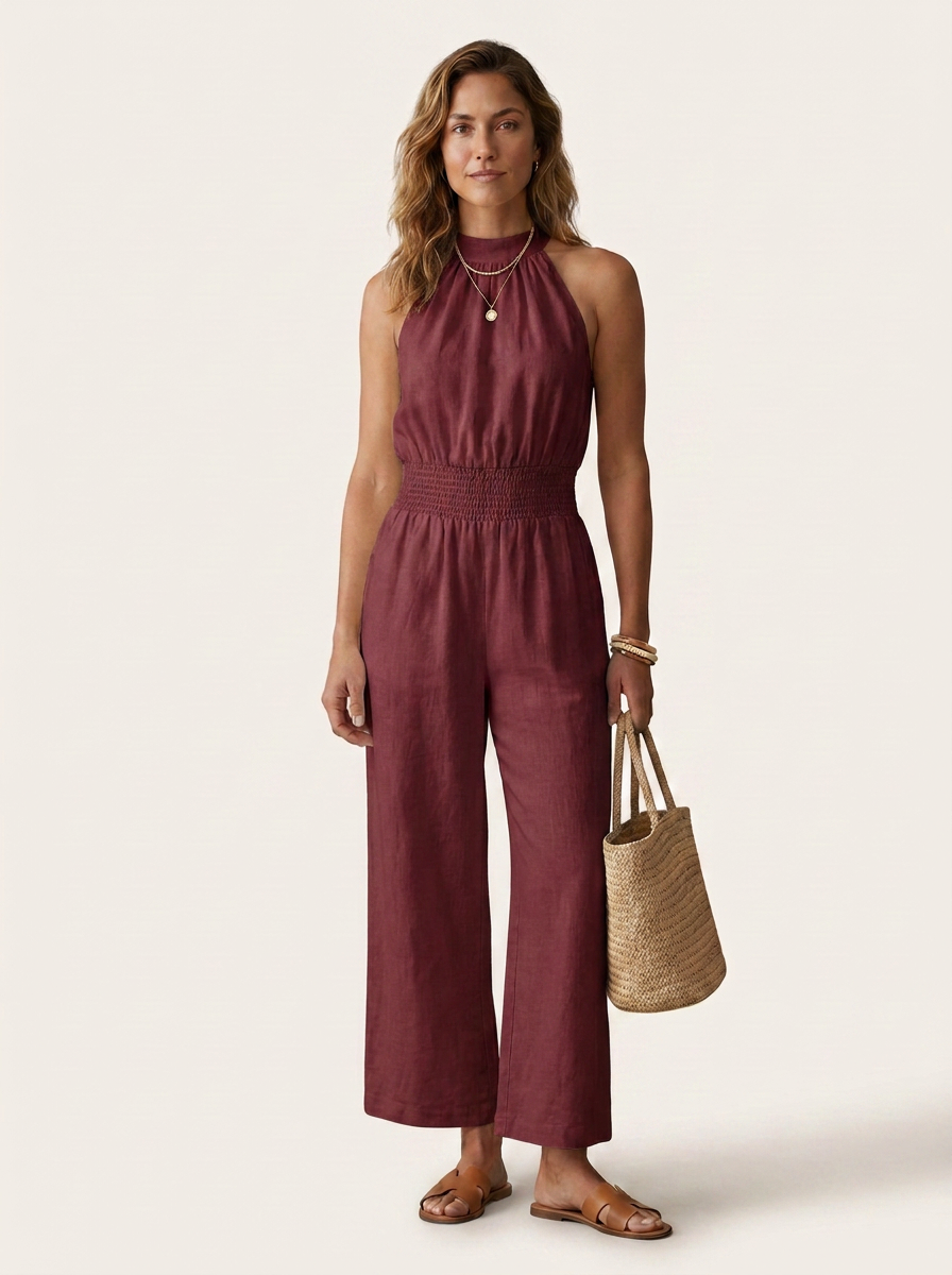 A woman modeling a sleeveless, high-neck, wide-leg jumpsuit in a deep burgundy or maroon shade. She is standing against a solid, warm off-white background and holding a woven straw tote bag.