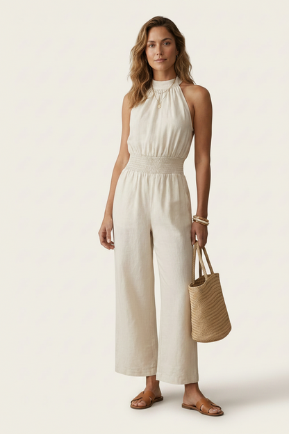 A woman models a sleeveless cream-colored wide-leg jumpsuit with a smocked waist, paired with brown flat sandals and a woven straw tote bag, set against a solid off-white studio background.