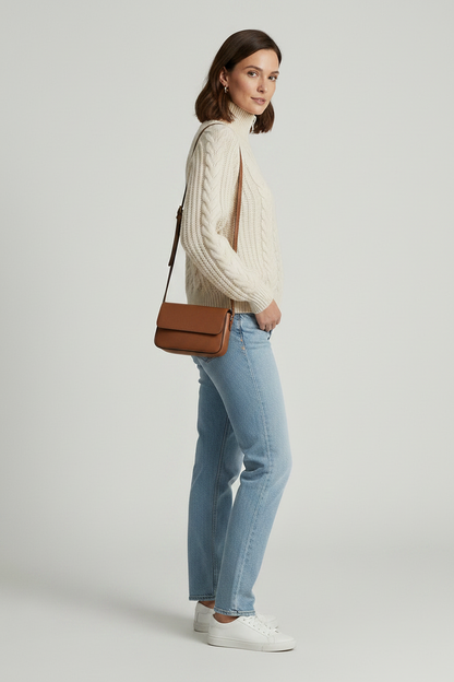 A woman stands against a light gray studio background wearing a cream cable-knit zip-front sweater, light blue jeans, white sneakers, and a small brown shoulder bag.