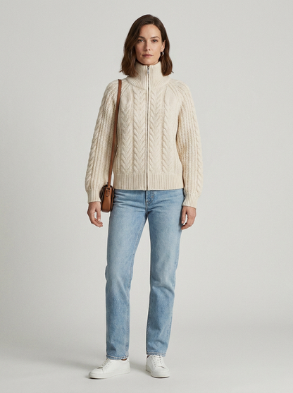 A woman stands against a light gray studio background wearing a cream cable-knit zip-front sweater, light blue jeans, white sneakers, and a small brown shoulder bag.