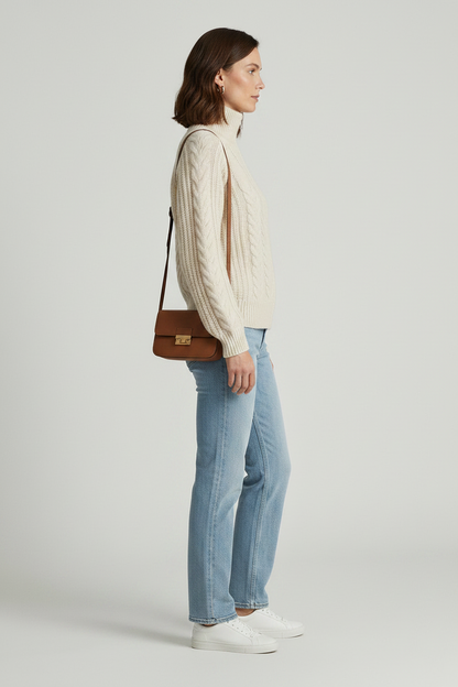 A woman modeling a cream cable-knit zip-up sweater with a mock neck, paired with light-wash blue jeans and white sneakers, against a light gray studio background.