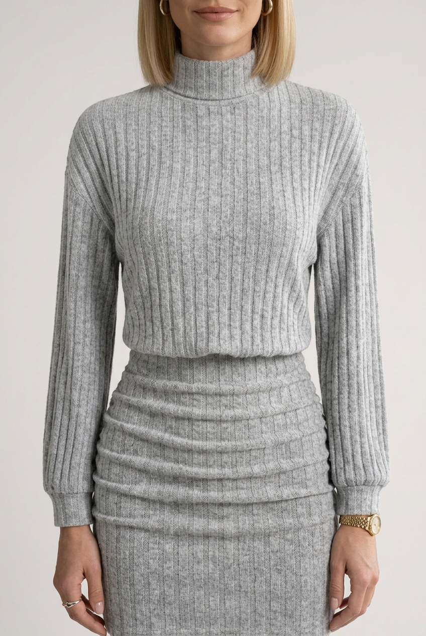 A woman models a light gray ribbed knit mock-neck long-sleeve top and matching pleated mini skirt set, styled with black ankle boots and a small black handbag against a plain light gray studio background.