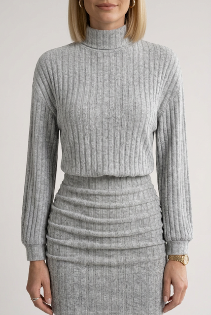 A woman models a light gray ribbed knit mock-neck long-sleeve top and matching pleated mini skirt set, styled with black ankle boots and a small black handbag against a plain light gray studio background.