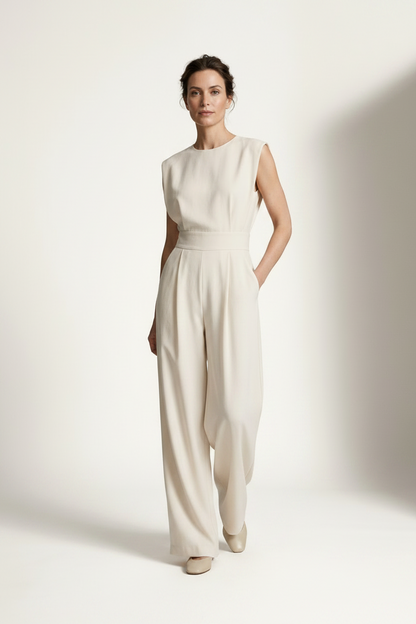 A woman modeling a sleeveless cream-colored wide-leg jumpsuit with a matching waistband, standing against a warm off-white studio background with soft shadows.