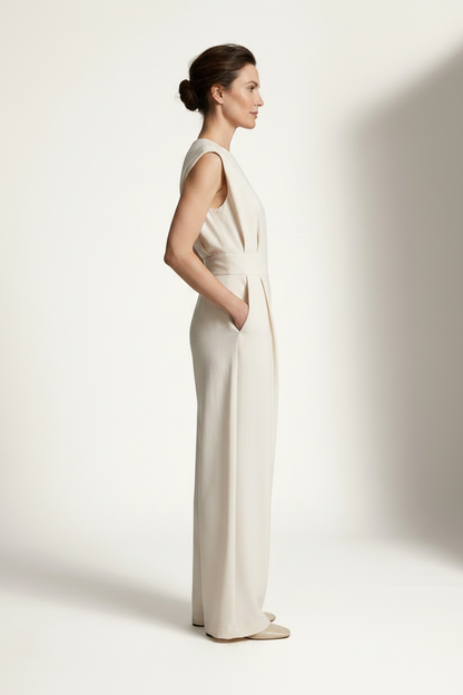 A woman modeling a sleeveless cream-colored wide-leg jumpsuit with a matching waistband, standing against a warm off-white studio background with soft shadows.