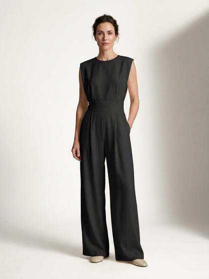 A woman modeling a black sleeveless wide-leg jumpsuit against a warm off-white studio background.