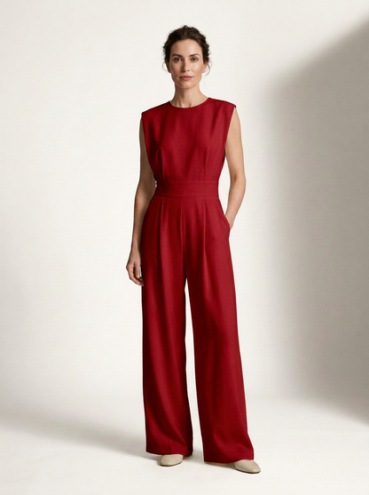 A woman modeling a sleeveless red jumpsuit with a defined waist and wide legs, standing against a plain off-white studio background.