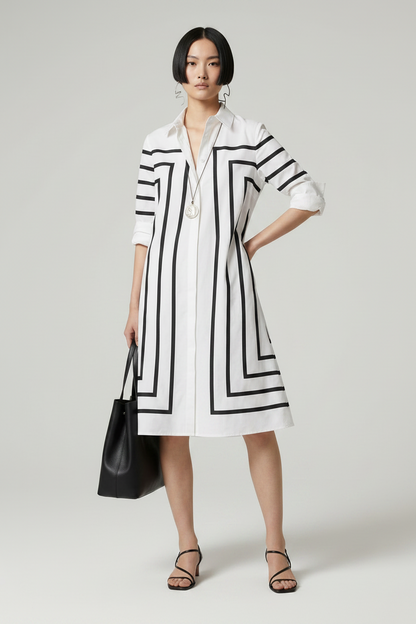 A model stands against a light gray studio background wearing a white long-sleeve shirt dress with bold black geometric stripe patterns. She is holding a black leather tote bag and wearing black strappy heels.
