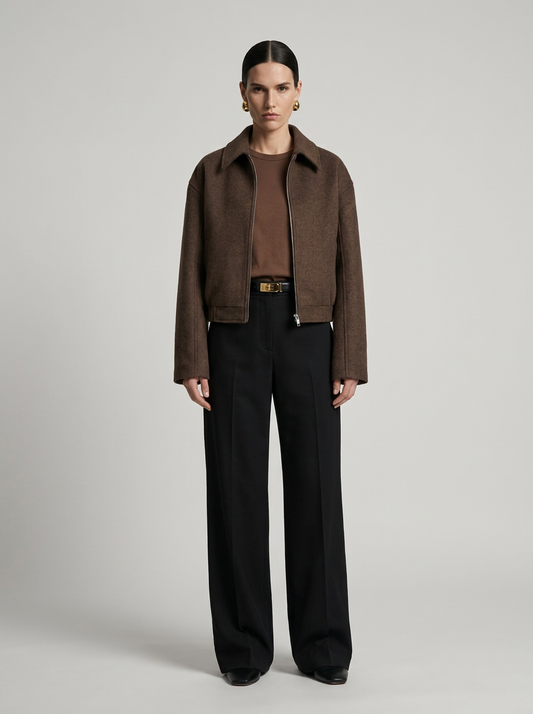 A model stands against a light gray studio background wearing a brown suede jacket over a brown top, black wide-leg trousers, and a black belt with a gold buckle.