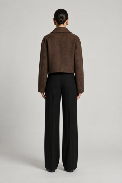 A model shown from the back wearing a cropped brown textured jacket and black wide-leg trousers, photographed against a neutral light gray studio background.