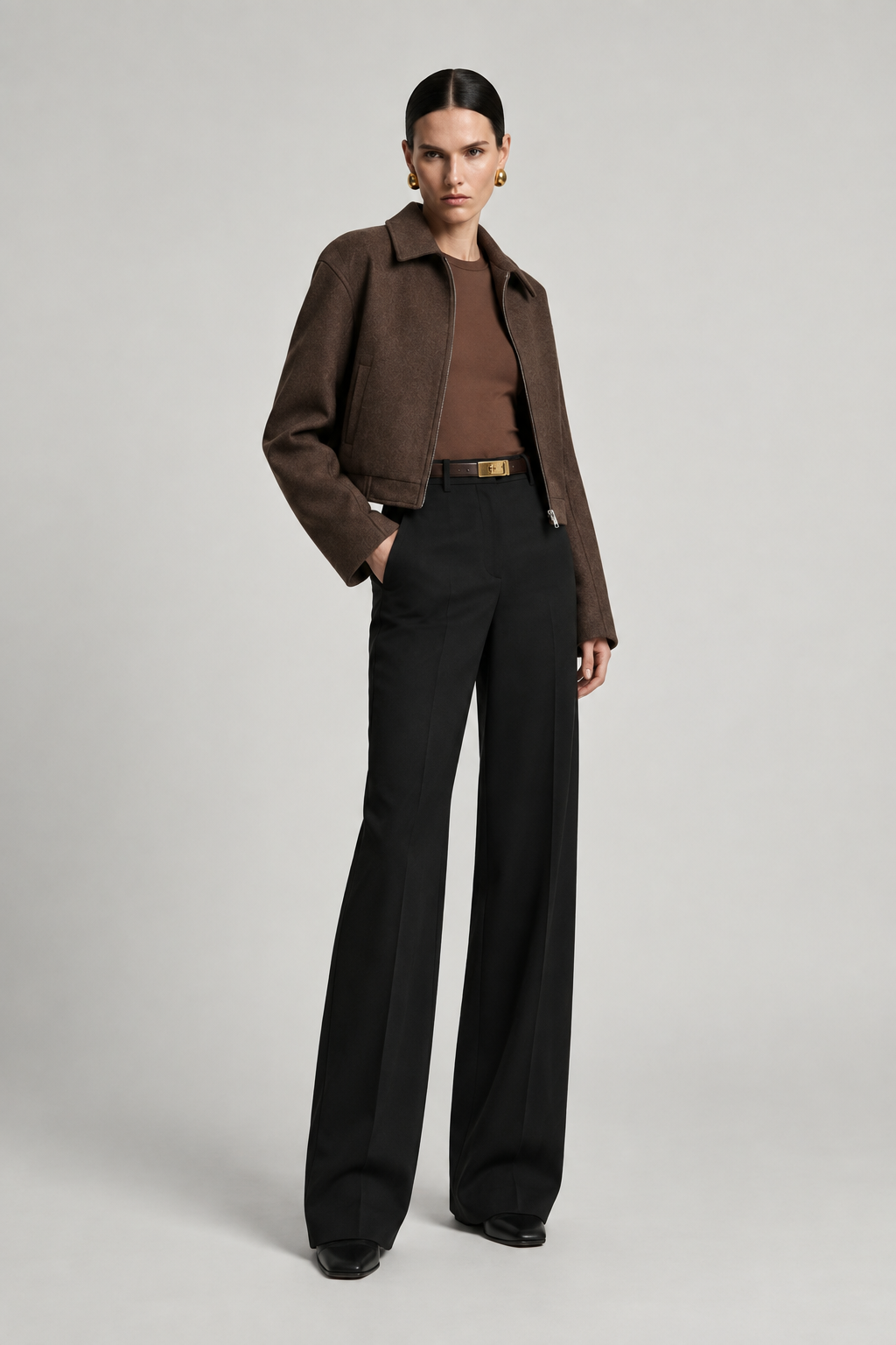 A model stands against a light gray studio background wearing a brown textured cropped jacket over a brown top, paired with black wide-leg trousers and black shoes.