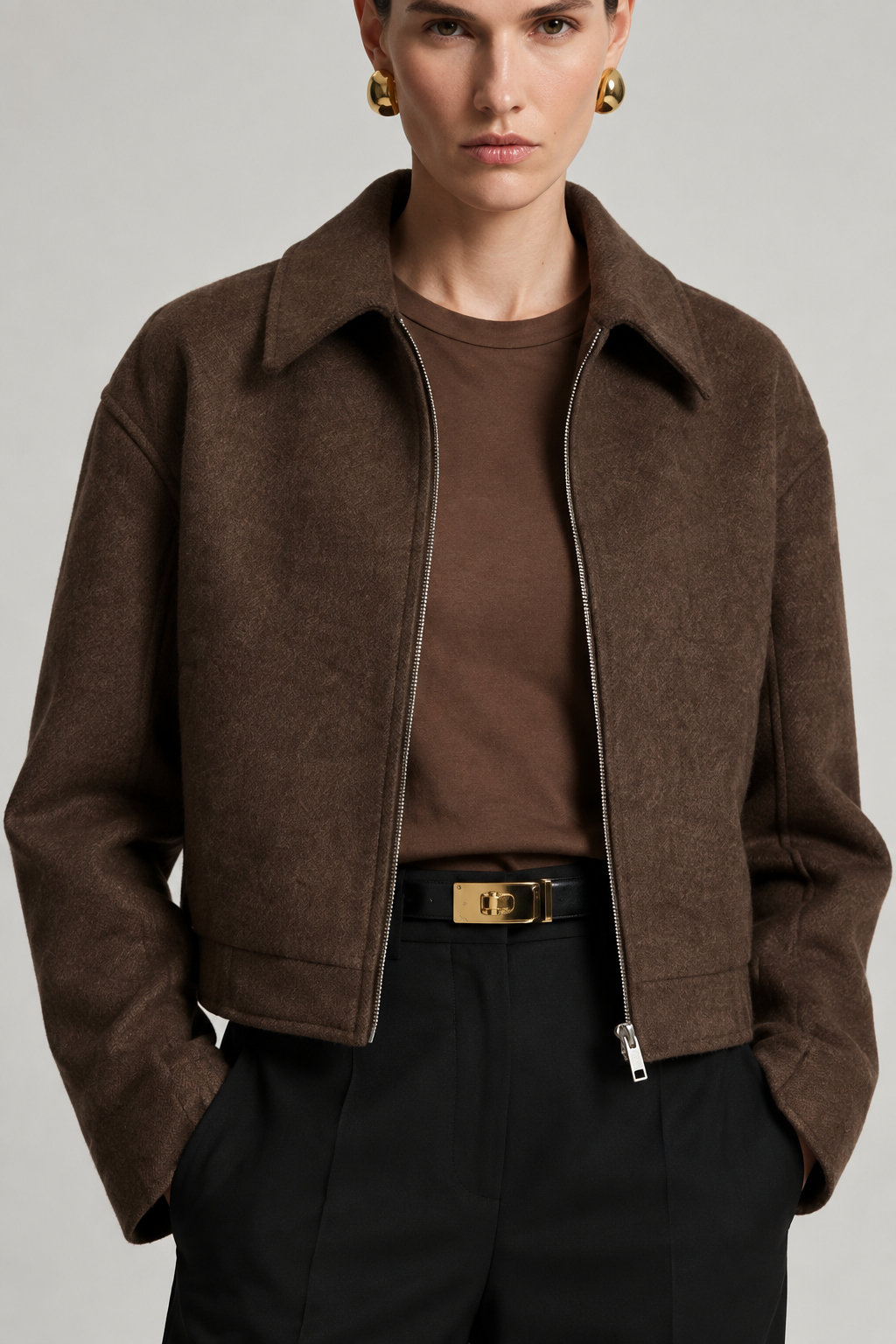 A model wearing a cropped brown suede jacket over a matching brown crewneck top, styled with black trousers and a black belt with a gold buckle.
