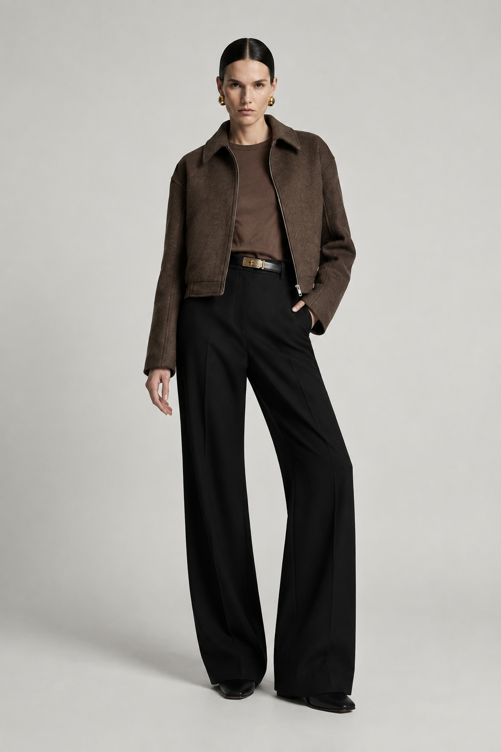 A model stands against a light gray studio background wearing a cropped brown suede jacket over a brown top, black wide-leg trousers, and black shoes.