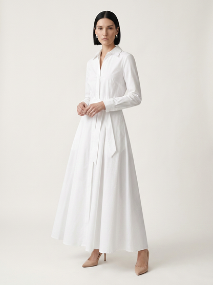A woman modeling a long-sleeve white shirt dress with a tie waist, standing against a plain light beige studio background.