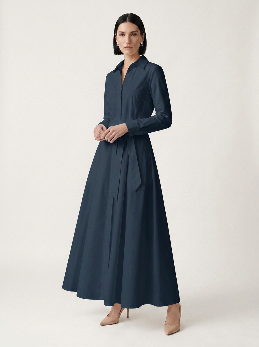 A woman modeling a long-sleeved navy blue maxi shirt dress with a tie waist, standing against a plain light beige studio background.