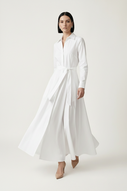 A woman modeling a long-sleeve white shirt dress with a tie waist, standing against a plain light beige studio background.