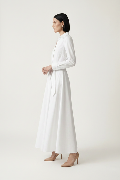 A woman modeling a long-sleeve white shirt dress with a tie waist, standing against a plain light beige studio background.